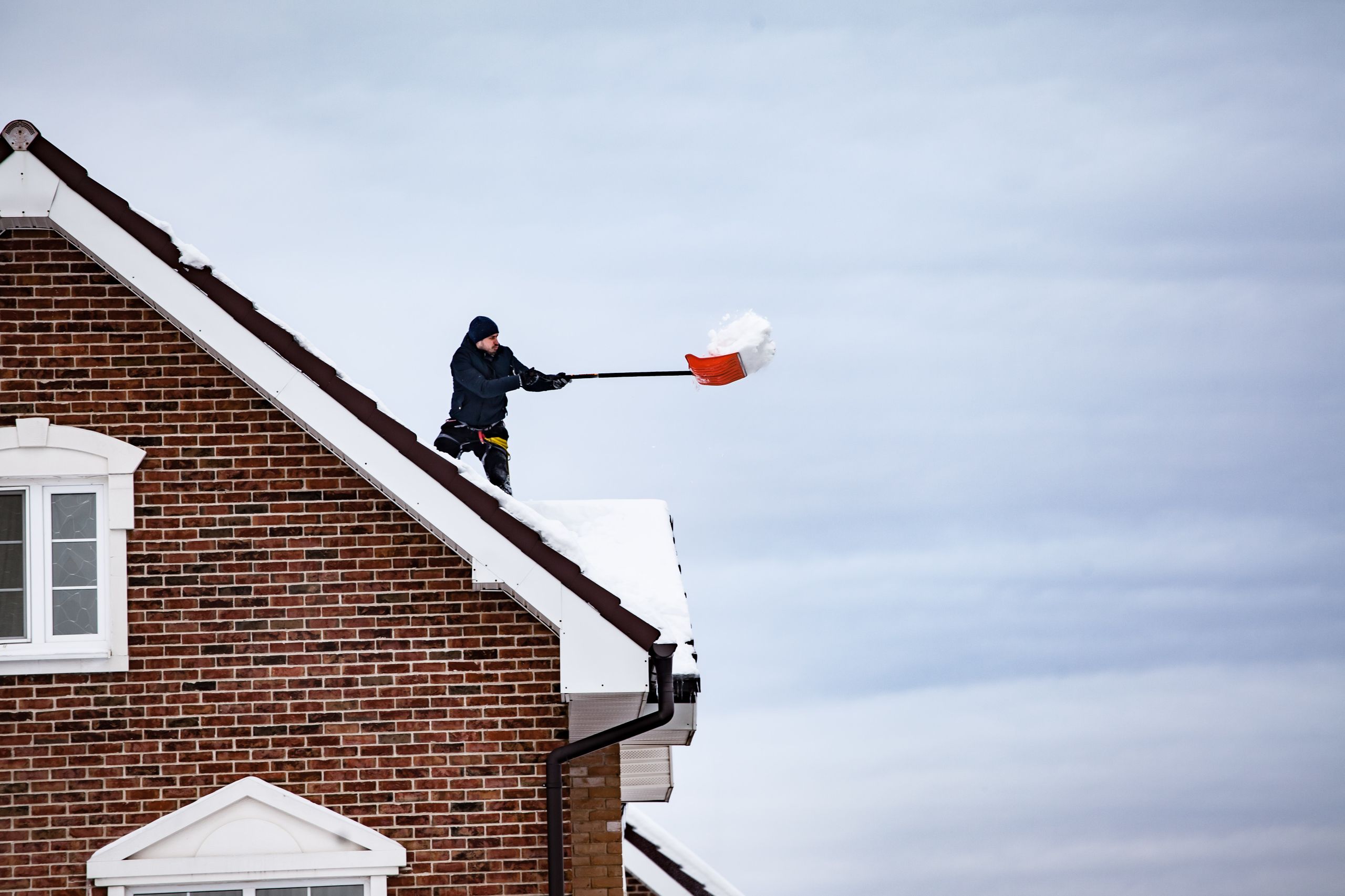A man shoveling snow off the roof of a brick house during winter weather.